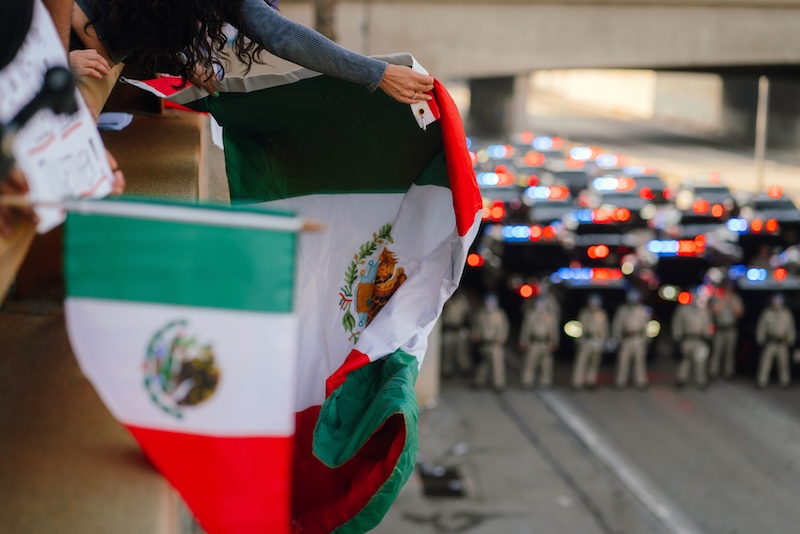 Several people hold the Mexican flag above a freeway filled with police vehicles and line of law enforcement officers on June 2025 in L.A. in protest of the rampant disappearance of immigrant and refugee communities by ICE.