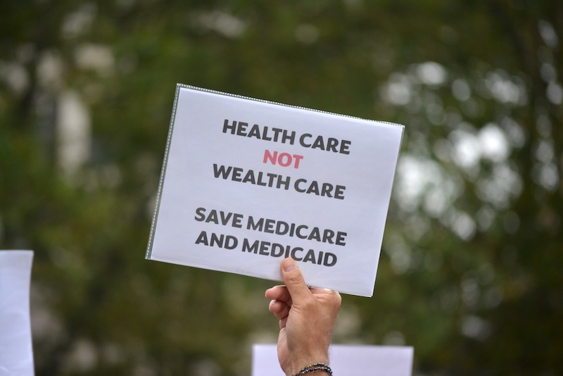 A hand holds a white sign with black and red letters. It says: Health Care Not Wealth Care, Save Medicare and Medicaid
